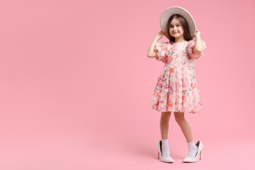 Stylish little girl wearing oversized high heeled shoes on pink background, space for text