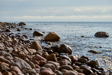 Rocky shoreline extends into tranquil ocean, with smooth stones scattered along coast. Gentle waves...