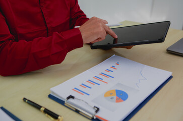 An Asian businessman in a red shirt sits thoughtfully at his desk, and analyzing charts, deep in...
