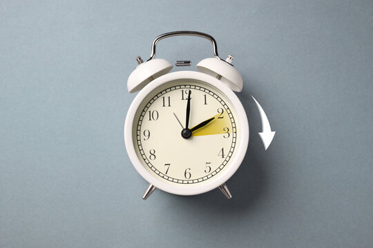 Daylight saving time. White alarm clock on grey background, top view. Highlighted area and arrow showing time change by one hour forward