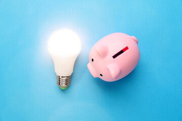 Piggy bank and glowing light bulb on blue background, top view. Energy saving concept
