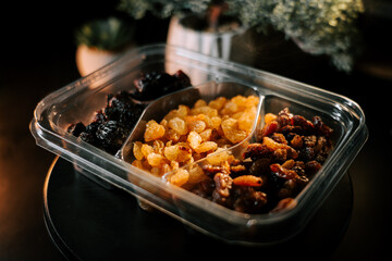 Variety of raisins and dried fruits in container displayed on dark surface with plant background