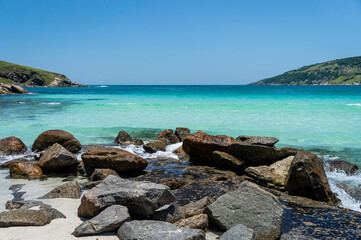 Rock formations at the northwest end of Prainha beach meeting Atlantic Ocean turquoise waters under bright and hot summer afternoon sunny clear blue sky in Arraial do Cabo, Rio de Janeiro - Brazil.