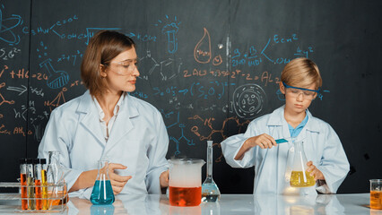 Caucasian teacher explained about colored solution at science laboratory. Expert instructor support smart boy doing experiment in STEM science class at blackboard written chemical theory. Erudition. © InfiniteFlow