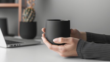 Two hands holding modern matte black coffee cup sitting at minimalist workstation with laptop in background.