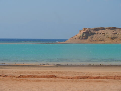 A stunning turquoise pool of water contrasts against a rugged, rocky shoreline and a clear blue sky in Saudi Arabia. The scene offers a serene and picturesque view of the Red Sea coast.