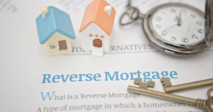 Homeownership, financial planning for retirees, and leveraging home equity for financial needs. Two small miniature house models, keys, and a pocket watch placed on a document titled Reverse Mortgage.