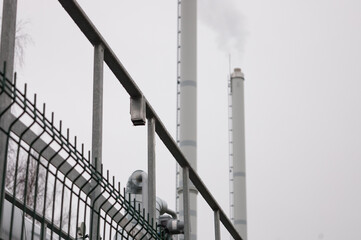 pipeline and pipeline elements behind a metal fence against the background of a gray sky and tall plant pipes