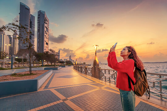 Female tourist taking pictures of modern skyscrapers at sunset from the Corniche promenade in Abu Dhabi, United Arab Emirates