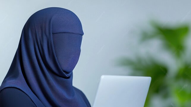 Muslim woman in a blue hijab and niqab uses a laptop computer while working in a modern office.