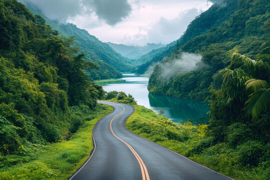 Winding through lush greenery, this scenic road leads to tranquil waters, surrounded by majestic mountains and misty skies, creating a perfect backdrop for a peaceful journey.