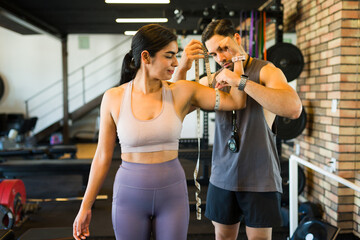Hispanic personal trainer measuring biceps of female client with measuring tape during fitness assessment at gym