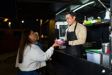 Cheerful cook presenting fresh tacos to customer near illuminated food truck, capturing street food excitement under evening lights