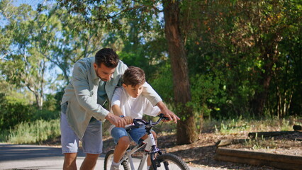 Father helping son ride bicycle in sunbeams park. Small child learning cycling