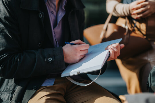 A person is taking handwritten notes in a notebook during an outdoor meeting. The scene conveys a sense of focus and engagement in a casual setting.
