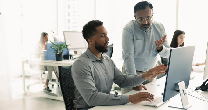 Senior Indian mentor checking work progress of African American intern man, looking at monitor of employee, speaking, giving consultation, advice. Young employee reporting to boss