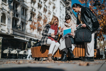 Three women dressed professionally discuss documents while sitting outdoors on a sunny day, depicting teamwork, learning, and collaboration.