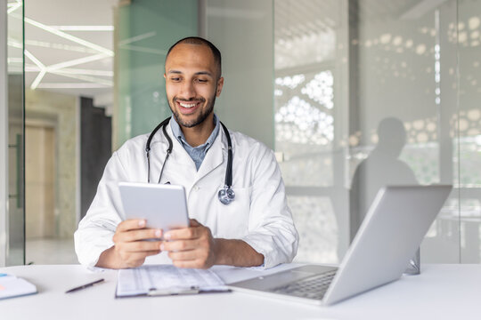 Successful experienced doctor sitting at desk inside the clinic office and using a tablet computer. Man in a white medical coat, using an application for online communication and consulting patients.