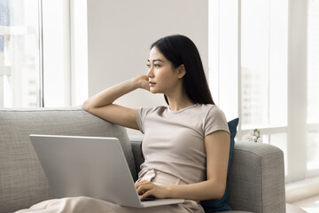Calm woman gazing thoughtfully into distance seated on sofa with laptop, in moment of deep...