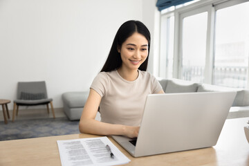 Young Asian woman working on her laptop in fashionable living room, smile, engaged in online tasks, studying or working. Creative, professional workflow using modern technology and internet connection