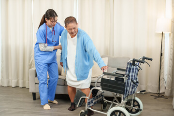 Physical therapist takes care elderly person after recovering from illness. Young therapist in uniform helps senior woman stand up from sofa while holding walker. Rehabilitation and elderly support