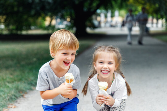 Naklejki Portrait two cute cheerful children enjoy ice cream together at nature city park path on hot summer sunny day. Funny kids eat sweet vanilla icecream walking family outdoors