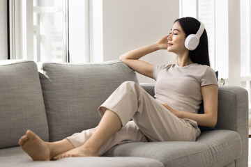 Obraz premium Young 25s Asian relaxed woman relish favourite music, wear modern wireless headphones, listening song or podcast with eyes closed, looking meditative and peaceful. Hobby time, weekend leisure at home