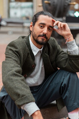 Handsome fashion stylish hipster guy in trendy casual clothes with green coat sitting on street in...