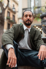 Naklejka premium Handsome spanish trendy man in fashion clothes with green coat sitting on street in Barcelona