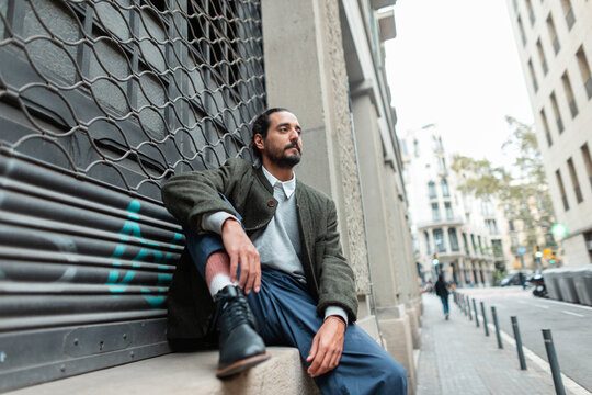 Stylish handsome fashion man in trendy clothes with green coat sitting on street near building in Barcelona city - Powered by Adobe