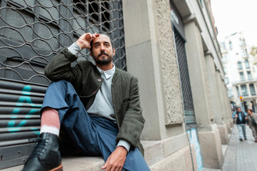 Handsome spanish trendy man in fashion clothes with green coat sitting on street in Barcelona