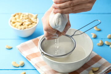 Woman straining cashew milk into bowl at light blue wooden table with nuts, closeup