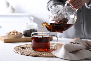 Woman pouring hot black tea into cup at white table, closeup