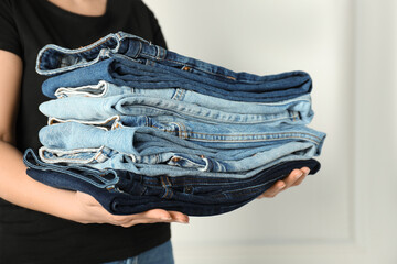 Woman with stack of different stylish jeans near light wall indoors, closeup
