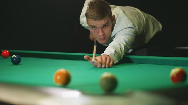 Close-up of focused player in white shirt leaning over green pool table, striking white billiard ball with cue stick. Intense gaze and precise aim capture skill, concentration in competitive billiards