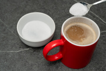 Hand holding spoon and is putting sugar into a red cup of coffee. Sugar crystals are trickling into...