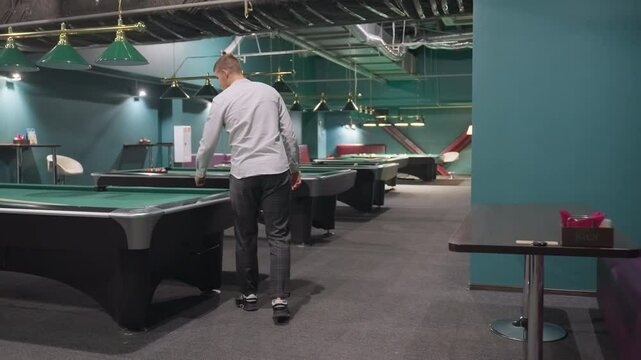ack shot of player in white shirt and grey trousers walking into billiard room, standing in front of green pool table, pick up white ball. Modern sports club setting with overhead lighting atmosphere