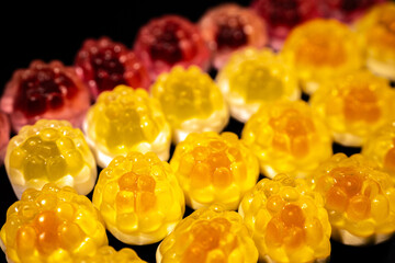 Red and yellow gummy candies resembling raspberries and pineapples are arranged on a black background. The candies have a shiny, translucent texture.
