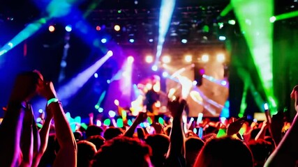 Slow motion captures the vibrant atmosphere of a concert, showcasing an enthusiastic crowd waving illuminated smartphones and glow sticks amidst colorful laser lights