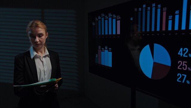 Young woman broker standing in office boardroom, analyzing financial data infographics on big display, holding folder with documents, checking information.