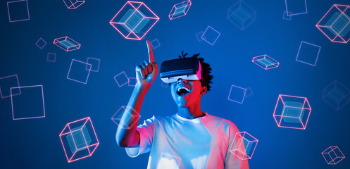 Smart boy standing at blue background while floating geometric shape. African man playing game and wearing VR goggle and casual shirt. Augmented reality technology innovation concept. Ingenuity.