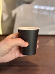 Unknown hand holding brown disposable paper cup with coffee from first face. Close-up