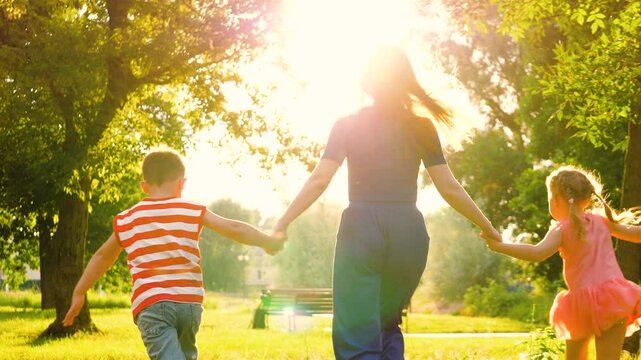 Silhouette of mother with hands interlocked with children leading across park lawn. Mother and children run across park with hands interlocked in bond of shared joy. Mother leads children through park