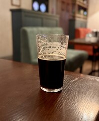 An unfinished transparent glass mug with dark beer and foam on the bar in an elite pub. Close-up