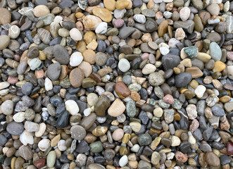 The image is a close-up of a pile of small pebbles or stones. The stones are of various sizes and colors, including shades of brown, beige, white, black, and gray. Some of the stones have a rough text