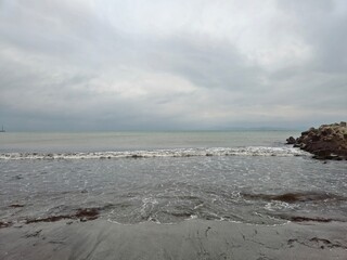 Wild waves roll onto a sandy beach beside large rocks, with a moody gray sky overhead
