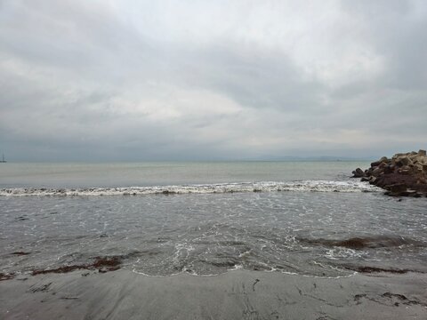 Wild waves roll onto a sandy beach beside large rocks, with a moody gray sky overhead