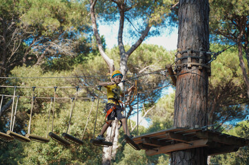 Naklejka premium Little boy in equipment and with insurance goes through obstacles at a height in a rope park in the forest. Extreme sport for children