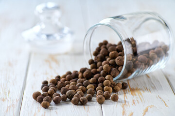 allspice or Jamaican pepper seeds spill out of a glass storage jar on a white wooden table, selective focus.