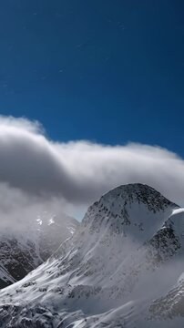 A stunning video capturing a snow-covered mountain peak with clouds flowing over it, set against a clear blue sky.
